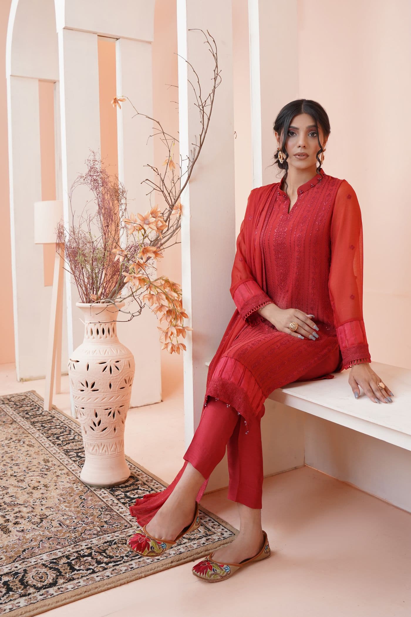 Woman in a red kurta and trousers seated on a bench in a peach-toned room